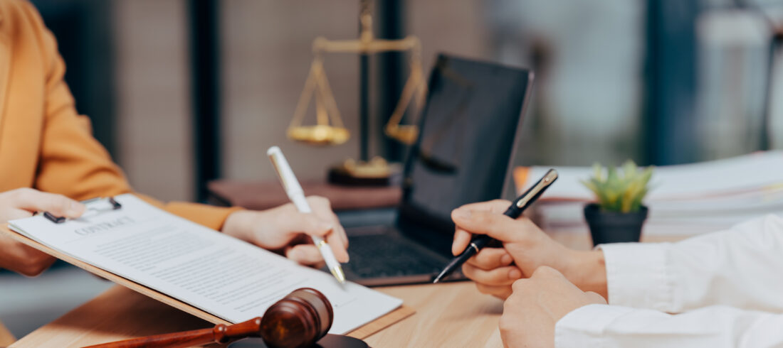 Legal Consultation: Two professionals sit at a desk, carefully reviewing and signing legal documents. In the background a scale of justice rests, and a gavel sits nearby.